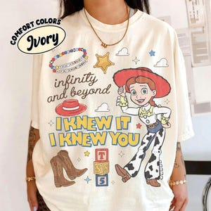 May include: An ivory t-shirt featuring a graphic print of the character Jessie from Toy Story, a red cowboy hat, brown cowboy boots, a beaded bracelet, and alphabet blocks. The text reads "infinity and beyond" and "I KNEW IT I KNEW YOU".