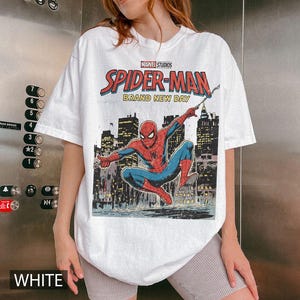 May include: White t-shirt featuring a Marvel Studios Spider-Man graphic. The design includes the text "SPIDER-MAN BRAND NEW DAY" with an illustration of Spider-Man in a cityscape setting. The shirt is a casual, short-sleeved style.