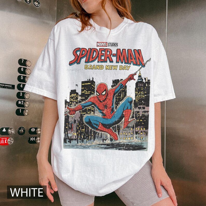 May include: White t-shirt featuring a Marvel Studios Spider-Man graphic. The design includes the text "SPIDER-MAN BRAND NEW DAY" with an illustration of Spider-Man in a cityscape setting. The shirt is a casual, short-sleeved style.