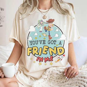 K&ouml;nnte beinhalten: Ein cremefarbenes Kurzarm-T-Shirt mit einer Grafik von Buzz Lightyear und Woody aus Toy Story. Das Design zeigt einen hellblauen Kreis, wei&szlig;e Wolken, gelbe Sterne, eine kleine Rakete und den Schriftzug YOU'VE GOT A FRIEND IN ME.