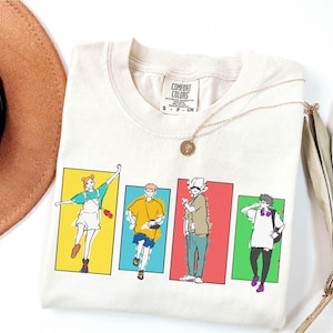 May include: Cream-colored t-shirt featuring a colorful graphic of four anime-style characters in rectangular frames. The shirt has a Comfort Colors tag and is accessorized with a gold necklace. A brown hat and a feather are partially visible.
