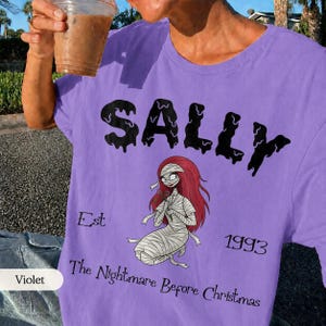 K&ouml;nnte beinhalten: Ein hellviolettes T-Shirt mit einer schwarzen Grafik der Figur Sally aus The Nightmare Before Christmas. Der Text lautet SALLY, Est 1993 und The Nightmare Before Christmas. Die Figur hat langes rotes Haar und ist in Bandagen gewickelt.