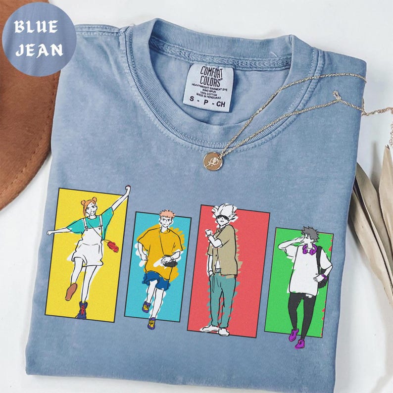 May include: A blue jean colored t-shirt with a graphic of four anime-style characters in colorful rectangular panels. The shirt has a crew neck and short sleeves. A gold necklace with a pendant is draped across the shirt.
