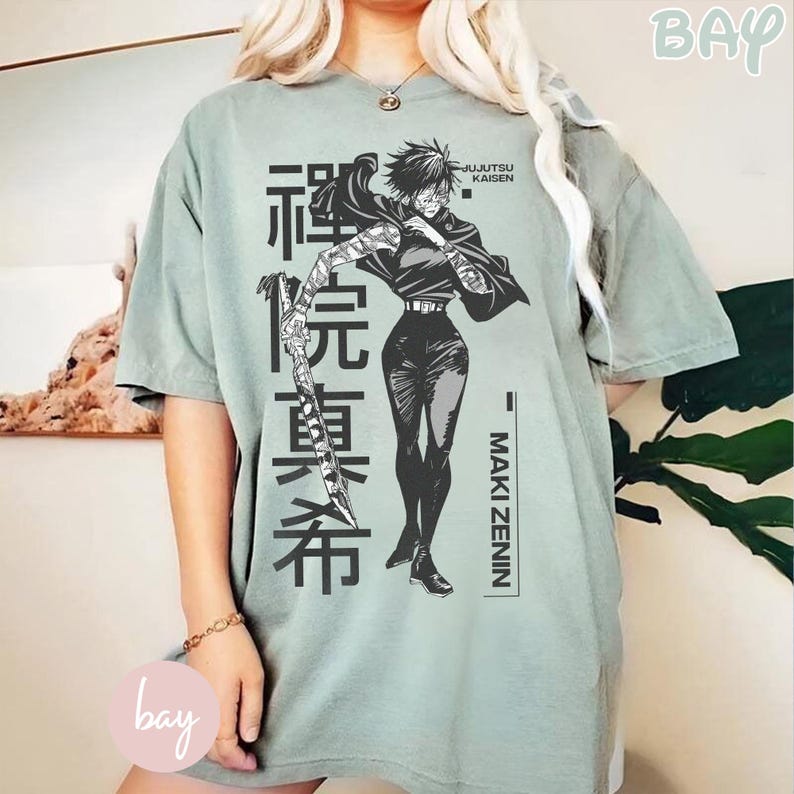 May include: Sage green t-shirt featuring a black and white illustration of Maki Zenin from Jujutsu Kaisen. The design includes Japanese text and the character's name. The shirt has short sleeves and a relaxed fit.