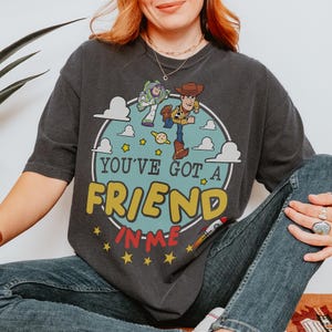 K&ouml;nnte beinhalten: Ein anthrazitfarbenes Kurzarm-T-Shirt mit einer kreisf&ouml;rmigen Grafik von Buzz Lightyear und Woody aus Toy Story. Das Design enth&auml;lt wei&szlig;e Wolken, gelbe Sterne, eine kleine Rakete und den Text "YOU'VE GOT A FRIEND IN ME" in Gelb und Rot.