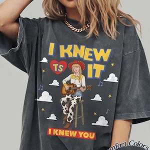 May include: A charcoal gray t-shirt featuring a graphic of a person in a cowboy hat, yellow shirt, and cow-print pants playing an acoustic guitar on a stool. The text reads I KNEW IT with a red heart containing TS and I KNEW YOU below, with clouds.