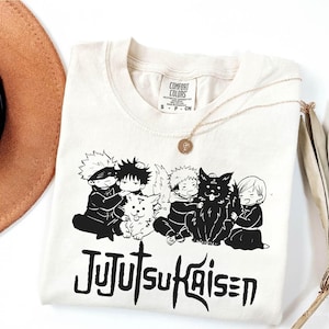 May include: Cream-colored t-shirt featuring a black graphic of anime characters and the text "Jujutsu Kaisen." The shirt has a crew neck and short sleeves. A gold necklace with a pendant is draped on the shirt.
