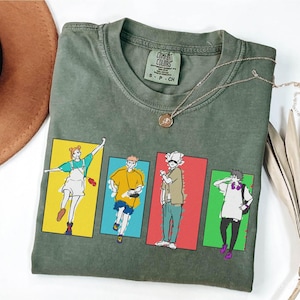 May include: Olive green t-shirt with a graphic of four anime characters in colorful rectangular frames. The shirt has a Comfort Colors tag and a gold necklace. The characters are in various poses, with distinct outfits and hairstyles.