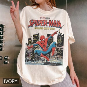 May include: Ivory-colored t-shirt featuring a Marvel Studios Spider-Man graphic. The design includes the text "SPIDER-MAN BRAND NEW DAY" above an illustration of Spider-Man in a cityscape setting. The shirt has short sleeves.