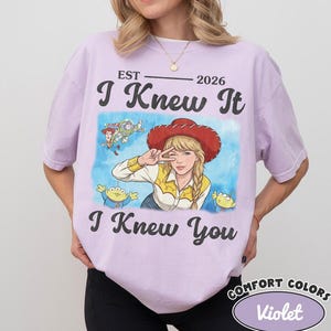 May include: A light purple short-sleeve t-shirt featuring a graphic print of a person wearing a red cowboy hat and yellow shirt, surrounded by cartoon characters. The text reads EST 2026, I Knew It, and I Knew You in black lettering.