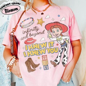May include: A light pink t-shirt featuring a graphic of a cowgirl character, a red hat, brown cowboy boots, a gold star, and alphabet blocks. The text reads infinity and beyond, I KNEW IT I KNEW YOU, and You've got a friend in me on a beaded bracelet.