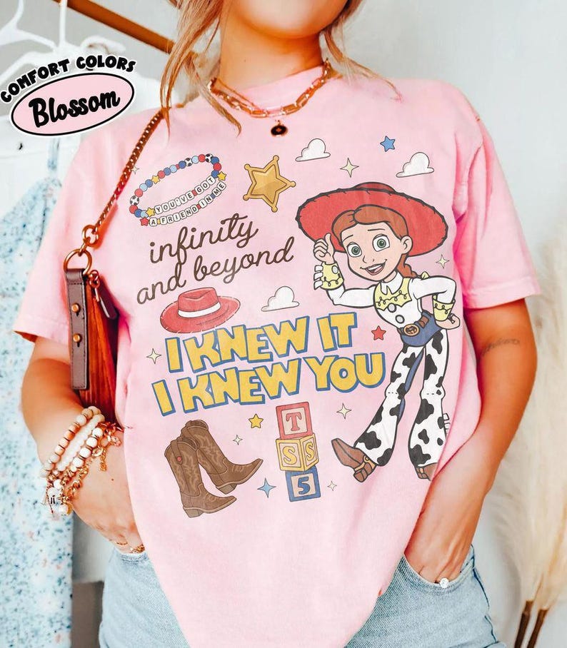 May include: A light pink t-shirt featuring a graphic of a cowgirl character, a red hat, brown cowboy boots, a gold star, and alphabet blocks. The text reads infinity and beyond, I KNEW IT I KNEW YOU, and You've got a friend in me on a beaded bracelet.