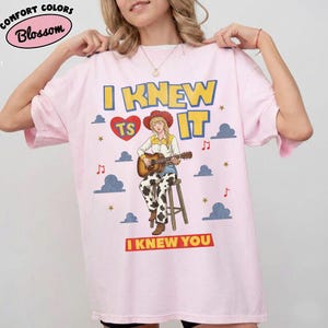 May include: A light pink t-shirt featuring a graphic of a person in a cowboy hat and cow-print pants playing an acoustic guitar while sitting on a stool. The text reads I KNEW TS IT and I KNEW YOU, surrounded by clouds, stars, and music notes.