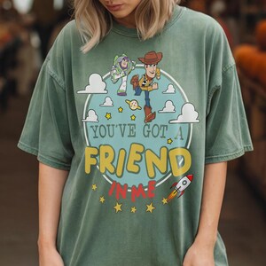 K&ouml;nnte beinhalten: Ein salbeigr&uuml;nes Kurzarm-T-Shirt mit einer Grafik von Woody und Buzz Lightyear. Das Design zeigt einen hellblauen Kreis, wei&szlig;e Wolken, gelbe Sterne, eine kleine Rakete und den Text "YOU'VE GOT A FRIEND IN ME" in Gelb und Rot.