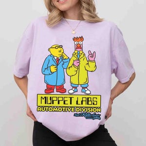 May include: A light purple short-sleeve t-shirt featuring a graphic of Dr. Bunsen Honeydew and Beaker. The text reads MUPPET LABS AUTOMOTIVE DIVISION and Rock 'n' Roller Coaster Starring Aerosmith. The characters are wearing lab coats.