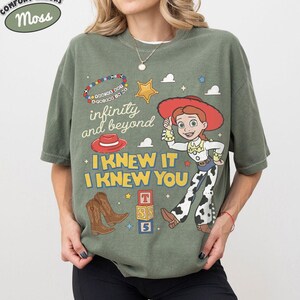 May include: An olive green t-shirt featuring a graphic print of a cowgirl character, brown cowboy boots, a red hat, and alphabet blocks. The text reads infinity and beyond, I KNEW IT, I KNEW YOU, and you've got a friend in me on a beaded bracelet.