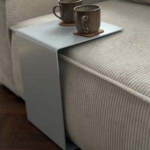 Puede incluir: Una mesa auxiliar en forma de C de color gris claro con un acabado liso y mate. La mesa presenta una superficie superior plana y un panel de soporte vertical que se desliza sobre el borde de un sofá. Dos tazas de cerámica marrón descansan sobre posavasos.