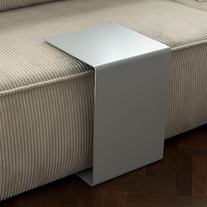 Può includere: Un tavolino a forma di C di colore grigio chiaro opaco con una superficie liscia e continua. Il tavolo presenta una sezione superiore piatta che si estende orizzontalmente e una base verticale che poggia sul pavimento accanto al divano.