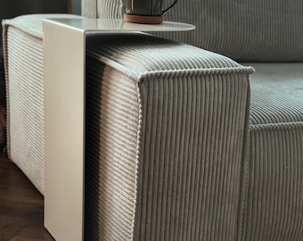 May include: A cream-colored, C-shaped metal side table with a flat, rectangular top and a vertical base that slides over the arm of a light gray, corduroy-textured sofa. A dark brown ceramic mug sits on the surface of the side table.