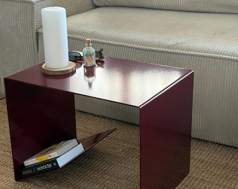 Puede incluir: Una mesa auxiliar de metal rojo oscuro con una superficie plana y lisa y un estante inclinado integrado debajo. La mesa sostiene una vela de pilar blanca sobre un posavasos de madera, un pequeño frasco de vidrio con cerillas y dos libros.