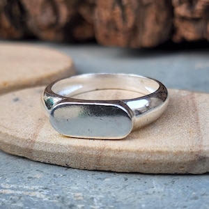 Könnte beinhalten: Ein polierter silberner Siegelring mit rechteckiger Oberfläche. Der Ring wird auf einem kleinen, ovalen, beigefarbenen Stein mit strukturiertem Holzgrund präsentiert. Das Design des Rings ist schlicht und klassisch.