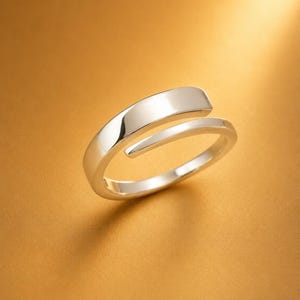 May include: A polished silver-tone bypass ring featuring a smooth, open-ended design with a flat, rectangular top section that tapers into a rounded band. The metal surface is reflective and captures light, showcasing a clean, continuous metallic form.