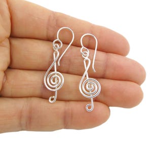 Könnte beinhalten: Ein Paar silberfarbene Ohrhänger in Form von Violinschlüssel-Symbolen. Jeder Ohrring besteht aus einem durchgehenden Drahtdesign, das die Spirale des Schlüssels bildet, und ist an einem einfachen, gebogenen Ohrhaken zur Befestigung aufgehängt.