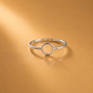 May include: A polished silver-toned ring featuring a thin, smooth band that connects to a centered, circular open-work element. The ring has a reflective metallic finish and is presented against a solid, warm-toned golden-brown background.