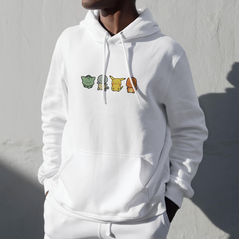 K&ouml;nnte beinhalten: Wei&szlig;er Kapuzenpullover mit einer Reihe von Cartoon-Figuren in Gr&uuml;n, Gelb und Orange. Der Pullover hat eine Vordertasche und eine Kapuze mit Kordelzug. Die Figuren sind horizontal &uuml;ber der Brust angeordnet.
