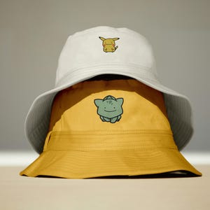 K&ouml;nnte beinhalten: Zwei &uuml;bereinander gestapelte Fischerh&uuml;te, einer wei&szlig; und einer gelb. Der wei&szlig;e Hut hat eine Pikachu-Stickerei, der gelbe einen Ditto. Beide H&uuml;te haben eine breite Krempe. Ein l&auml;ssiges Accessoire.