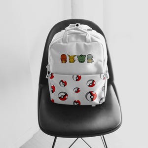 K&ouml;nnte beinhalten: Wei&szlig;er Rucksack mit einem Muster aus roten, schwarzen und wei&szlig;en Pok&eacute;-B&auml;llen auf der unteren H&auml;lfte. Die obere H&auml;lfte zeigt Cartoon-Figuren. Der Rucksack steht auf einem schwarzen Stuhl.