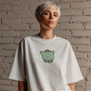 K&ouml;nnte beinhalten: Wei&szlig;es T-Shirt mit einem gr&uuml;nen Cartoon-Charakter-Motiv. Das T-Shirt hat kurze &Auml;rmel und einen Rundhalsausschnitt. Die Grafik ist ein gr&uuml;ner Cartoon-Charakter mit einem l&auml;chelnden Gesicht. Der Hintergrund ist eine wei&szlig;e Backsteinmauer.