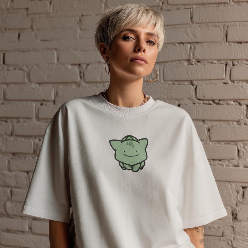K&ouml;nnte beinhalten: Wei&szlig;es T-Shirt mit einem gr&uuml;nen Cartoon-Charakter-Motiv. Das T-Shirt hat kurze &Auml;rmel und einen Rundhalsausschnitt. Die Grafik ist ein gr&uuml;ner Cartoon-Charakter mit einem l&auml;chelnden Gesicht. Der Hintergrund ist eine wei&szlig;e Backsteinmauer.