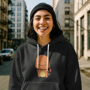 K&ouml;nnte beinhalten: Schwarzer Kapuzenpullover mit einem Charmander-Cartoon-Design in Orange, Gelb und Rot. Der Pullover hat eine Kapuze mit Kordelzug und eine Vordertasche. Die Person auf dem Bild tr&auml;gt eine schwarze M&uuml;tze.