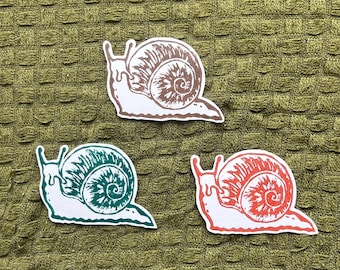 May include: Three die-cut stickers featuring snail illustrations. One snail is brown, one is dark green, and one is bright red. Each snail has a white body and a detailed, textured shell design with a spiral pattern, presented as individual decals.