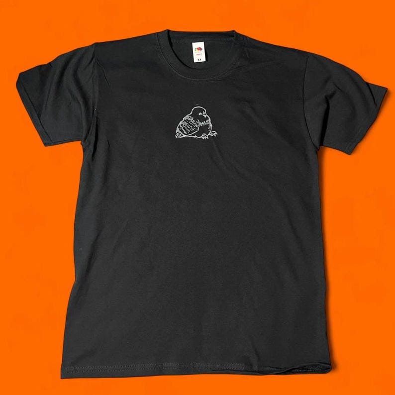 May include: Black t-shirt with a small, white, hand-drawn illustration of a bird on the front. The bird is depicted in a simple, minimalist style. The shirt is made of a soft material and has short sleeves.