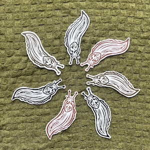 May include: Six die-cut stickers arranged in a circle, each featuring a stylized linocut illustration of a slug. The slugs are rendered in various solid colors including black, dark brown, and reddish-brown, all with white outlines and detailed line work.