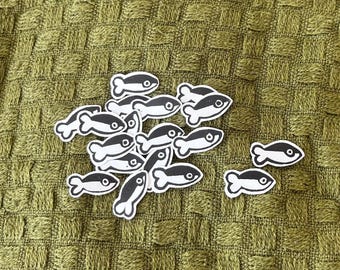May include: A collection of small, flat, die-cut paper shapes in the form of fish. Each fish shape features a black body with a white eye and a white outline. The fish are arranged in a cluster, with two separate fish shapes positioned to the right.