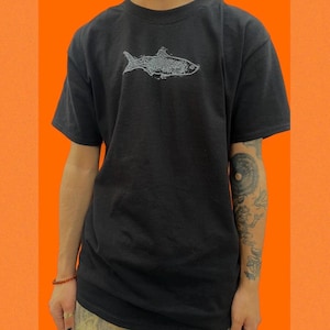 Peut inclure: T-shirt noir à manches courtes avec un motif de poisson blanc détaillé sur le devant. Le dessin du poisson est centré et semble être dessiné à la main. Le t-shirt est en matière souple.