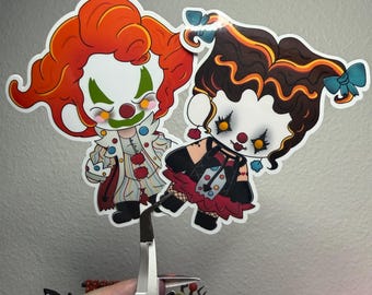 May include: Two die-cut stickers featuring stylized clown characters. The left character has bright orange hair, white face paint, and a striped outfit. The right character has dark brown hair with blue bows, white face paint, and a black and red dress.