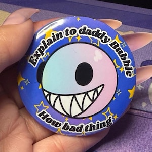 Puede incluir: Un botón redondo con fondo azul y el texto "Explain to daddy Bubble How bad thing". El botón presenta una cara de dibujos animados con un degradado de azul y rosa, una gran sonrisa y un ojo negro.