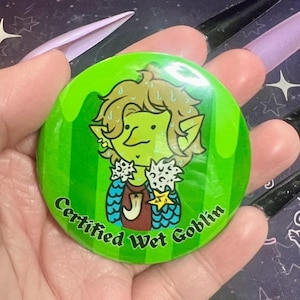 May include: A round, green button with a cartoon goblin illustration. The goblin has green skin, blonde hair, and is wearing a red shirt and blue and white accents. The text "Certified Wet Goblin" is printed around the edge.