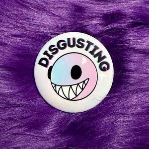 May include: A round white button with the word "DISGUSTING" in black and pink lettering. The button features a cartoon face with a black eye, a black mouth, and a row of white teeth. The face is split into two colors, blue and pink.