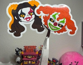 May include: Two die-cut vinyl stickers featuring stylized clown faces. One sticker depicts a character with black hair, yellow eyes, and red face paint. The other sticker shows a character with orange hair, green face paint, and a red nose.