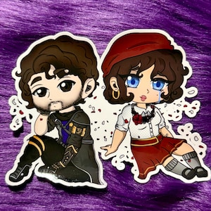 Könnte beinhalten: Zwei Aufkleber im Chibi-Stil, die eine männliche und eine weibliche Figur zeigen. Die männliche Figur trägt eine schwarze und goldene Rüstung, während die weibliche Figur ein rotes Barett und einen Rock trägt. Beide haben große Augen und sind von dekorativen Elementen umgeben.