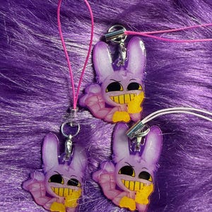 Puede incluir: Tres colgantes de conejito morado con dientes amarillos y una expresión sonriente, sujetos a cordones rosas y blancos. Los colgantes tienen un diseño de dibujos animados y están colocados sobre un fondo morado y difuso.