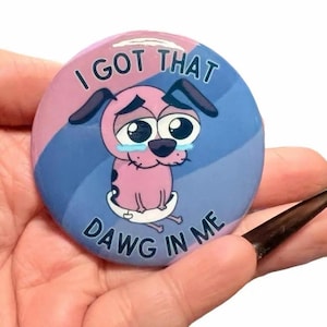 Puede incluir: Un botón redondo con una imagen de dibujos animados de Coraje, el perro cobarde, con el texto "I GOT THAT DAWG IN ME". El botón tiene un fondo rosa y azul con el perro en el centro, que parece triste con lágrimas.