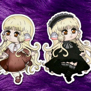 Puede incluir: Dos pegatinas con ilustraciones de estilo anime de un personaje parecido a una muñeca con cabello rubio y ojos marrones. Una muñeca lleva un vestido marrón con cuello blanco, la otra un vestido negro con una diadema negra. Ambas tienen un contorno blanco.
