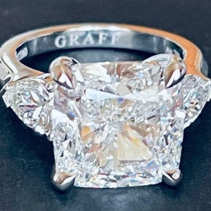 Puede incluir: Un anillo Graff con un gran diamante central de talla cuadrada, flanqueado por dos diamantes triangulares más pequeños. La banda plateada tiene la inscripción "GRAFF" en el interior.