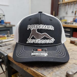 May include: Black and white trucker hat with the words "POTBELLIED STALLION" in white lettering. The hat features a brown graphic of a reclining figure. The hat has a white mesh back and a curved brim.